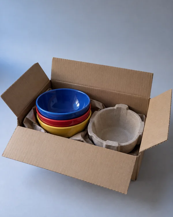 An open brown cardboard carton sits on a pale grey surface, holding three nested ceramic bowls with cobalt blue, red and yellow rims.