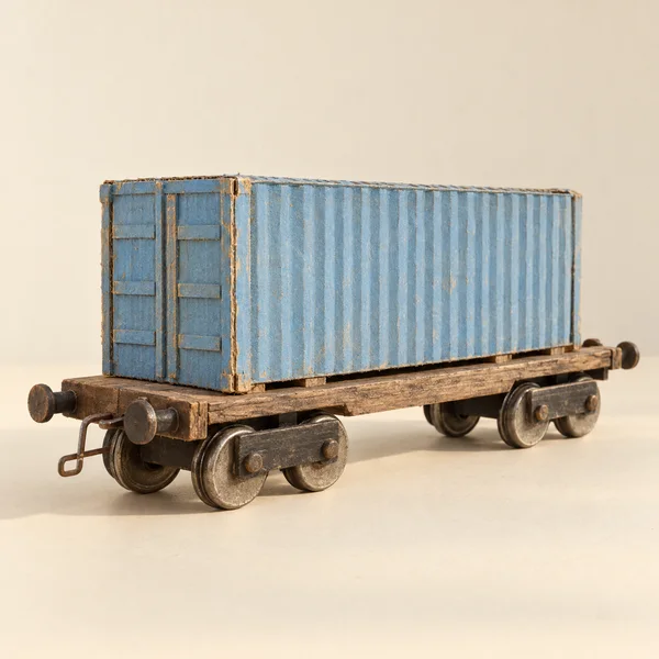 A weathered miniature flat wagon carries a faded blue corrugated container on a cream tabletop against a warm ivory background, with metal wheels, buffer ends…