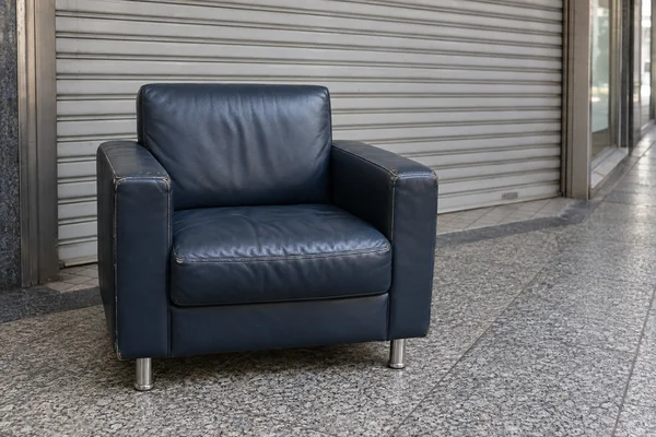 A navy-vinyl armchair with broad padded arms and chrome legs stands on gray terrazzo pavement in front of a closed metal roller shutter; shop glazing and a…
