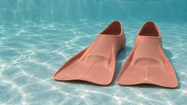 A matched pair of dusty-coral swim fins rests on a pale aqua tiled floor underwater, with their open foot pockets facing away and small bubbles inside.