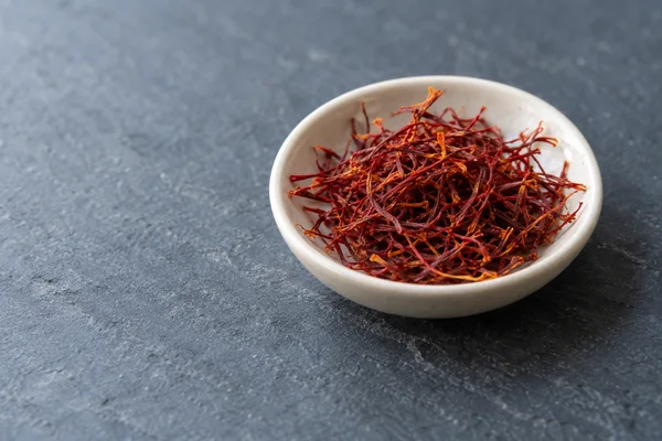 A shallow ivory porcelain dish filled with tangled crimson and orange saffron threads rests on a blue-gray slate surface, with open space to the left.