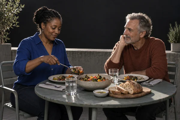 A woman in a cobalt shirt serves roasted vegetables at a round outdoor table while a gray-haired man in a rust sweater listens with his head resting on one…