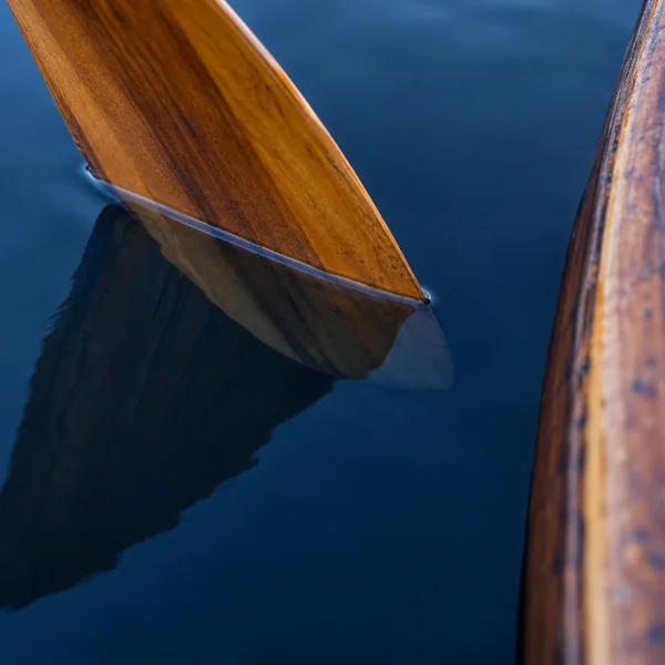 A warm wooden paddle blade angles down into still, deep blue water beside a curved wooden canoe hull at the right edge; the blade and its dark reflection meet…
