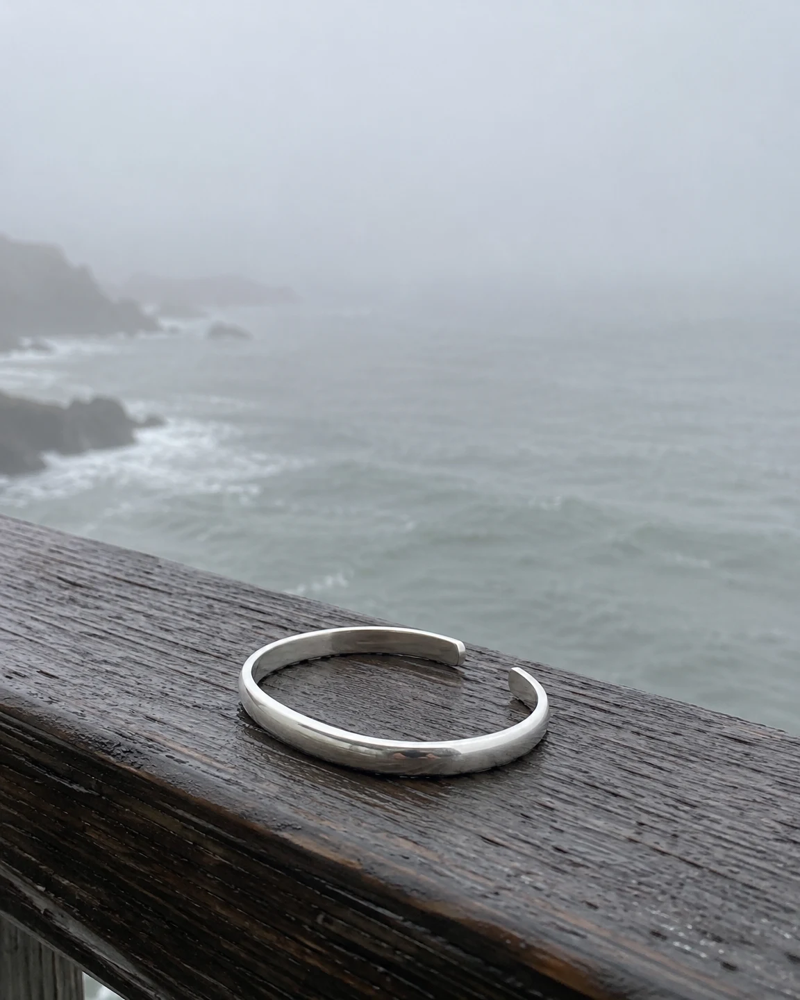 A plain silver open cuff bracelet lies on a damp, dark timber pier railing above a foggy grey sea, with a rocky shoreline faintly visible at left.