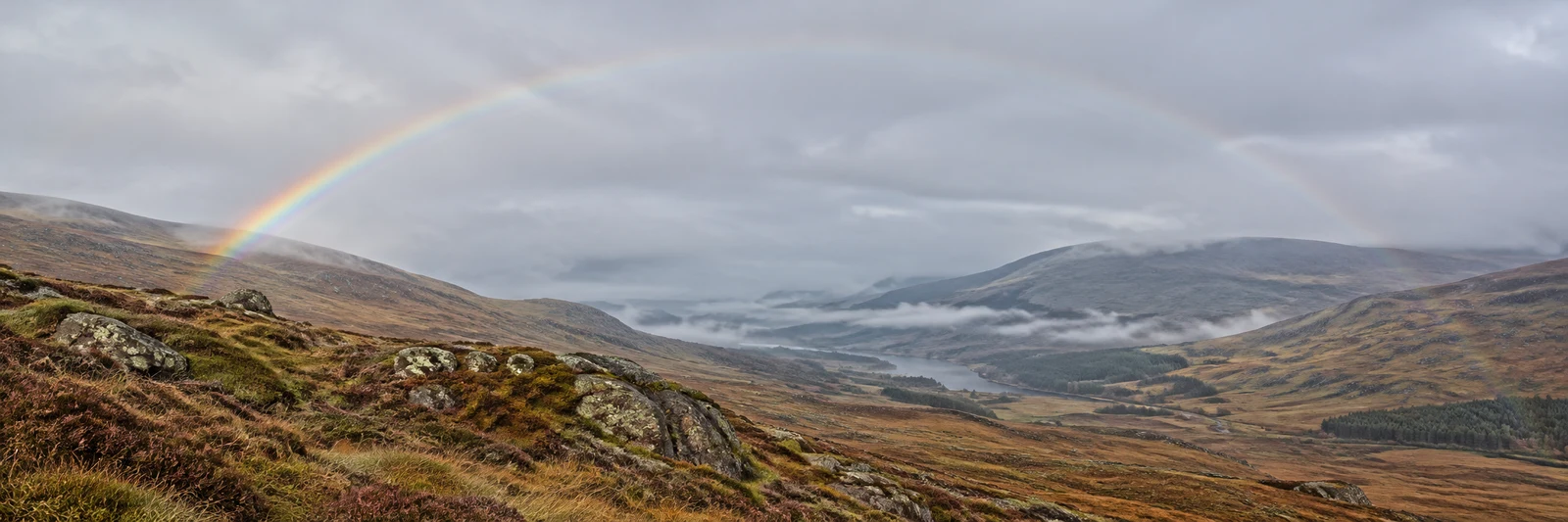 A broad moorland valley with rocky heather-covered slopes, low mist along the valley floor and a rainbow arching across a cloudy grey sky.