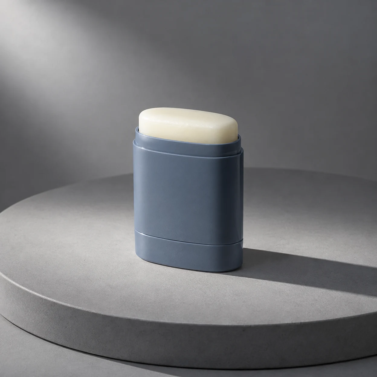 An uncapped slate-blue sunscreen stick with a raised ivory bar stands on a circular grey studio platform, lit from the upper left and casting a long shadow to…