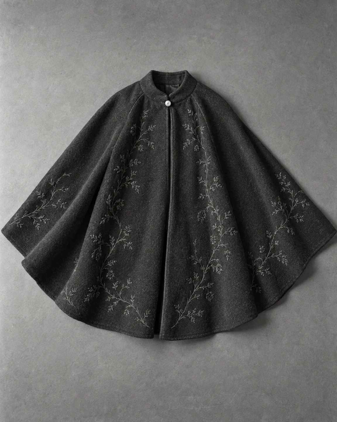 A charcoal wool cape with a rounded fan-shaped silhouette lies on a gray surface, showing a small light button at the neckline and branching embroidery across…