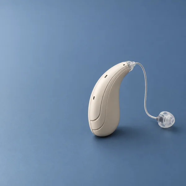 A warm ivory behind-the-ear hearing aid with recessed casing tiers rests on a matte slate-blue surface.