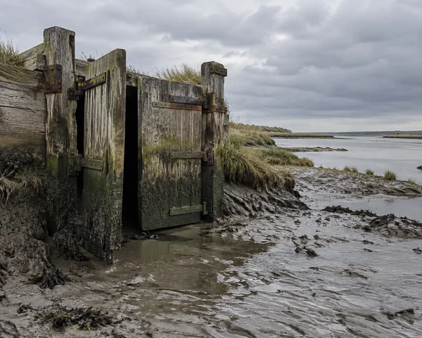 A weathered timber tidal sluice stands in a muddy estuary creek, with one gate leaf swung open beside a closed, algae-streaked leaf; saltmarsh grasses and a…