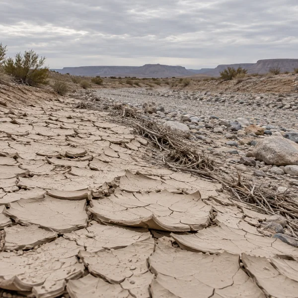 A dry desert wash with large upturned plates of cracked pale clay in the foreground, leading diagonally toward a gravel channel strewn with rounded stones and…
