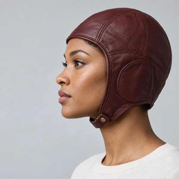 A woman with medium-brown skin wears a burgundy stitched leather aviator cap in left-facing profile against a pale gray studio background; a white ribbed…