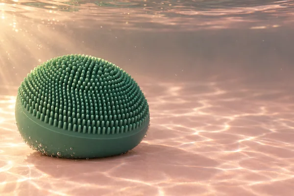 A domed dark emerald-green facial-cleansing brush rests on a pale blush floor underwater, with rows of short silicone nubs across its upper surface and small…