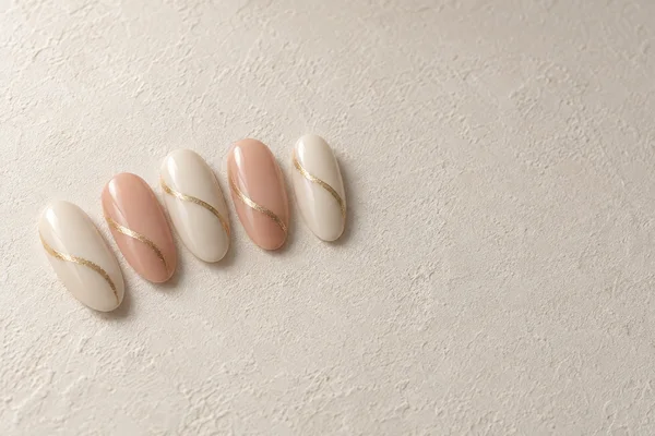 Five glossy press-on nail tips in alternating ivory and muted peach, each with a curved gold accent, are arranged in a loose fan on a textured ivory surface…