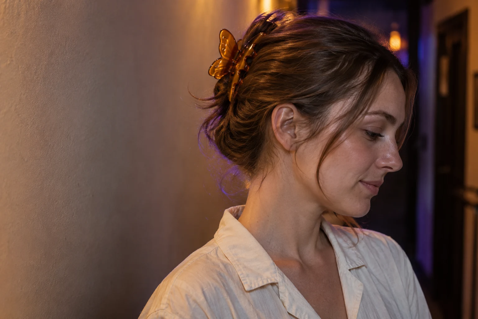 A woman in an off-white shirt pauses in profile in a dim venue corridor, looking slightly downward.