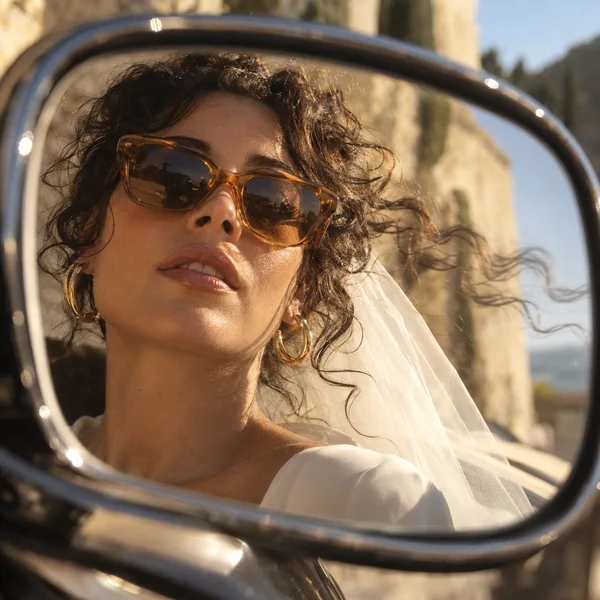 Close-up reflection of a bride with tousled dark curls, amber sunglasses and gold hoop earrings in a dark chrome side mirror, with an ivory veil and warm…