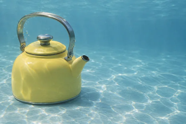 A butter-yellow enamel kettle with a stainless-steel arched handle and open spout sits on a pale blue underwater floor, with light patterns across the floor…