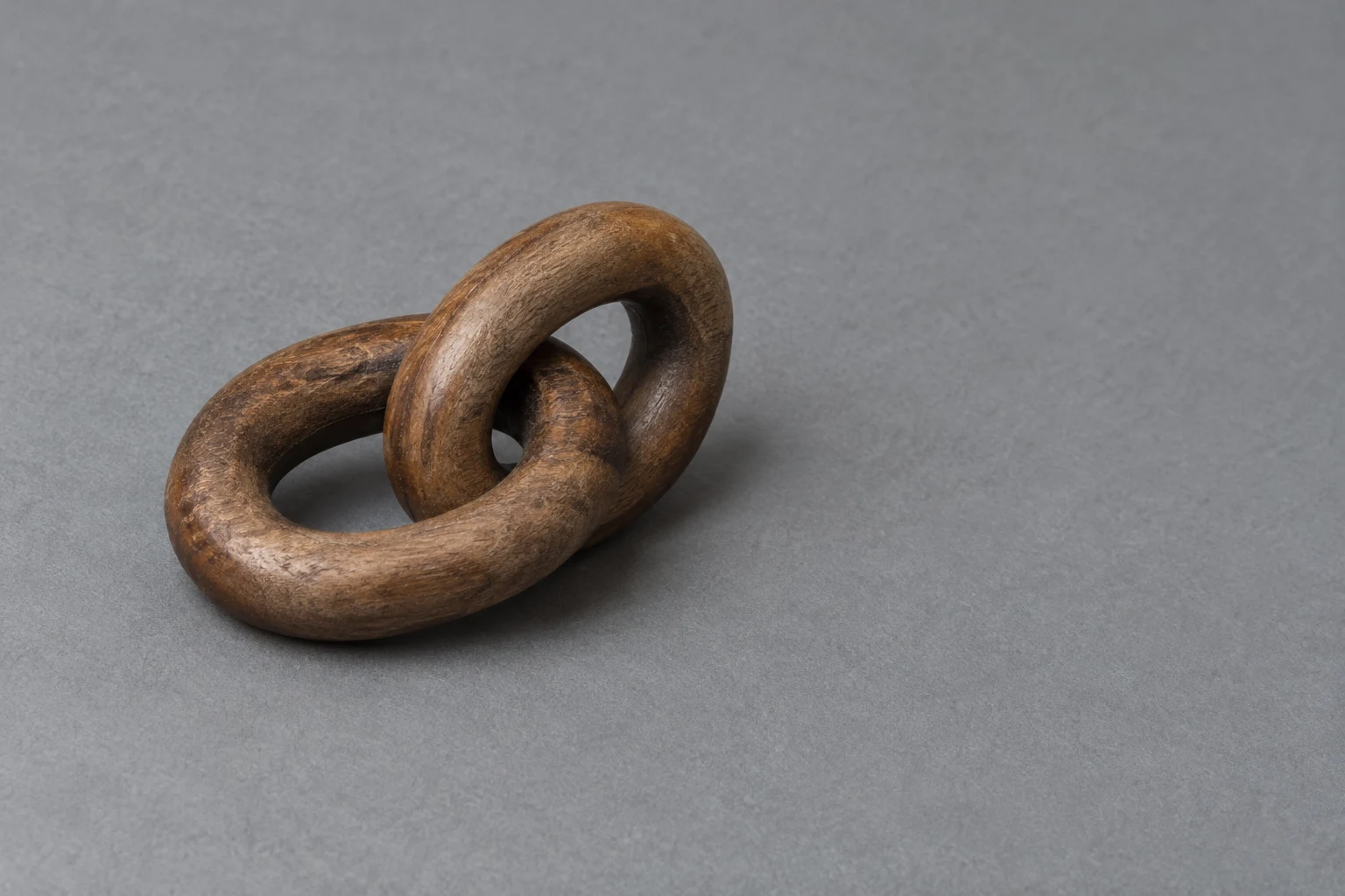 Two thick, brown wooden oval links interlock on a cool-gray textured mat, with one lying low and the other rising upright; the object sits left of center with…