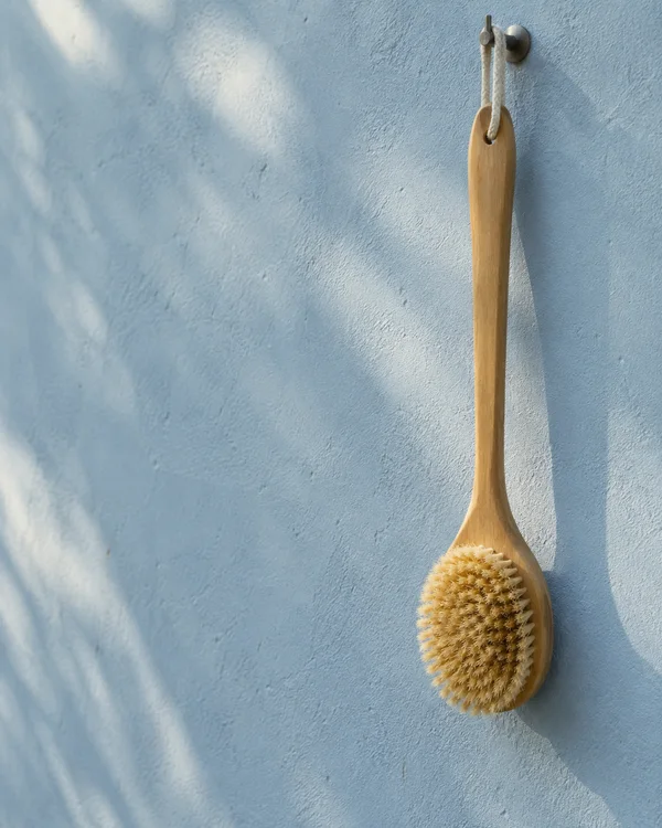 A long wooden bath brush hangs by a pale loop from a metal hook on a textured powder-blue wall, with its honey-colored bristles facing outward and open wall…