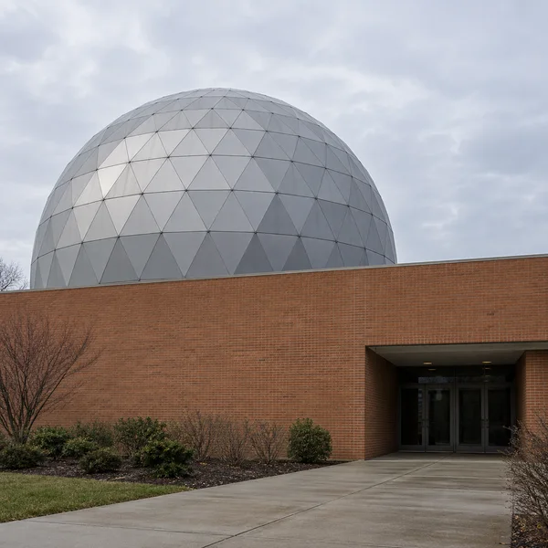 A silver hemispherical dome made of triangular panels rises behind a long terracotta brick wall, with a recessed glass entrance at the lower right under a pale…