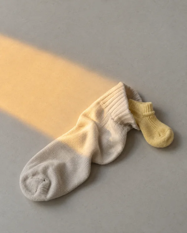 A rumpled cream adult sock lies on a pale grey tabletop with a small butter-yellow sock partly tucked beside its opening; a soft amber light patch crosses the…