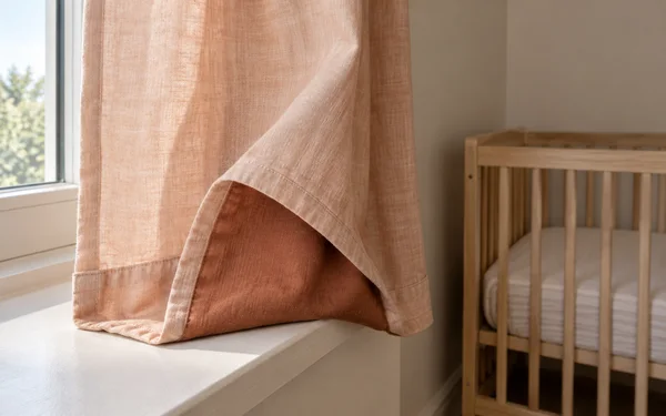 A linen nursery curtain drapes over a white windowsill, with one bottom corner turned back to reveal a darker terracotta inner hem.