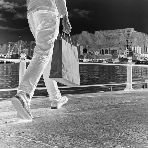 A waist-down view of a person in light trousers and sneakers walking beside a harbour railing, carrying a paper shopping bag; water, boats, buildings and a…