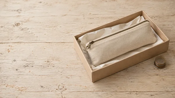 A natural-canvas zippered pencil case lies diagonally on ivory tissue inside a shallow kraft box on a pale, worn wooden tabletop, with a small stack of coins…
