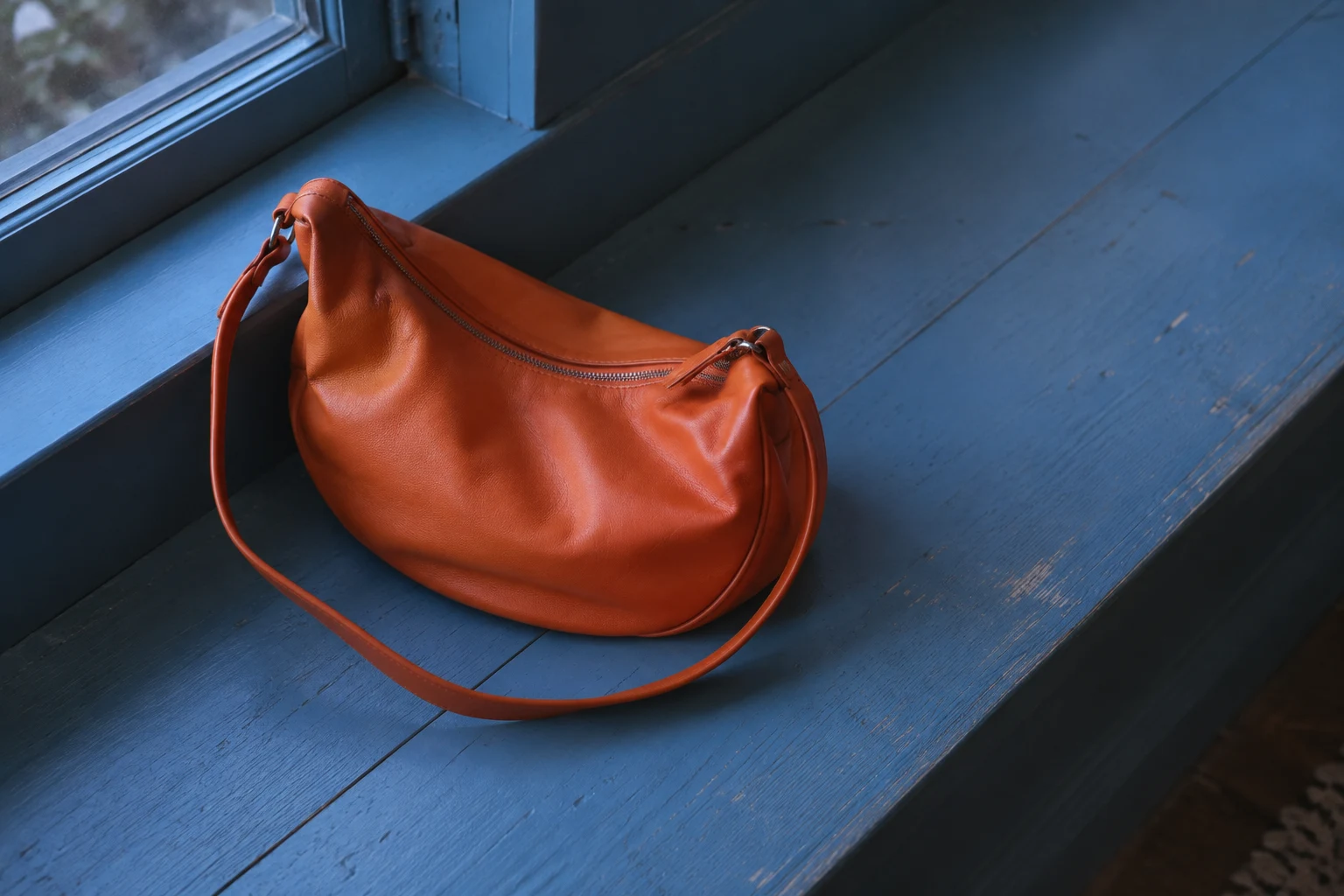 A tangerine-orange crescent handbag with a closed zipper rests on a blue-painted wooden window bench, its long strap looping across the seat; the bench and…