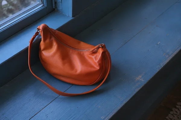 A tangerine-orange crescent handbag with a closed zipper rests on a blue-painted wooden window bench, its long strap looping across the seat; the bench and…