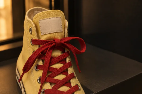 A butter-yellow canvas high-top sneaker with a blank cream tongue label and tomato-red laces sits on a charcoal shelf, warmly lit beside a glowing display…