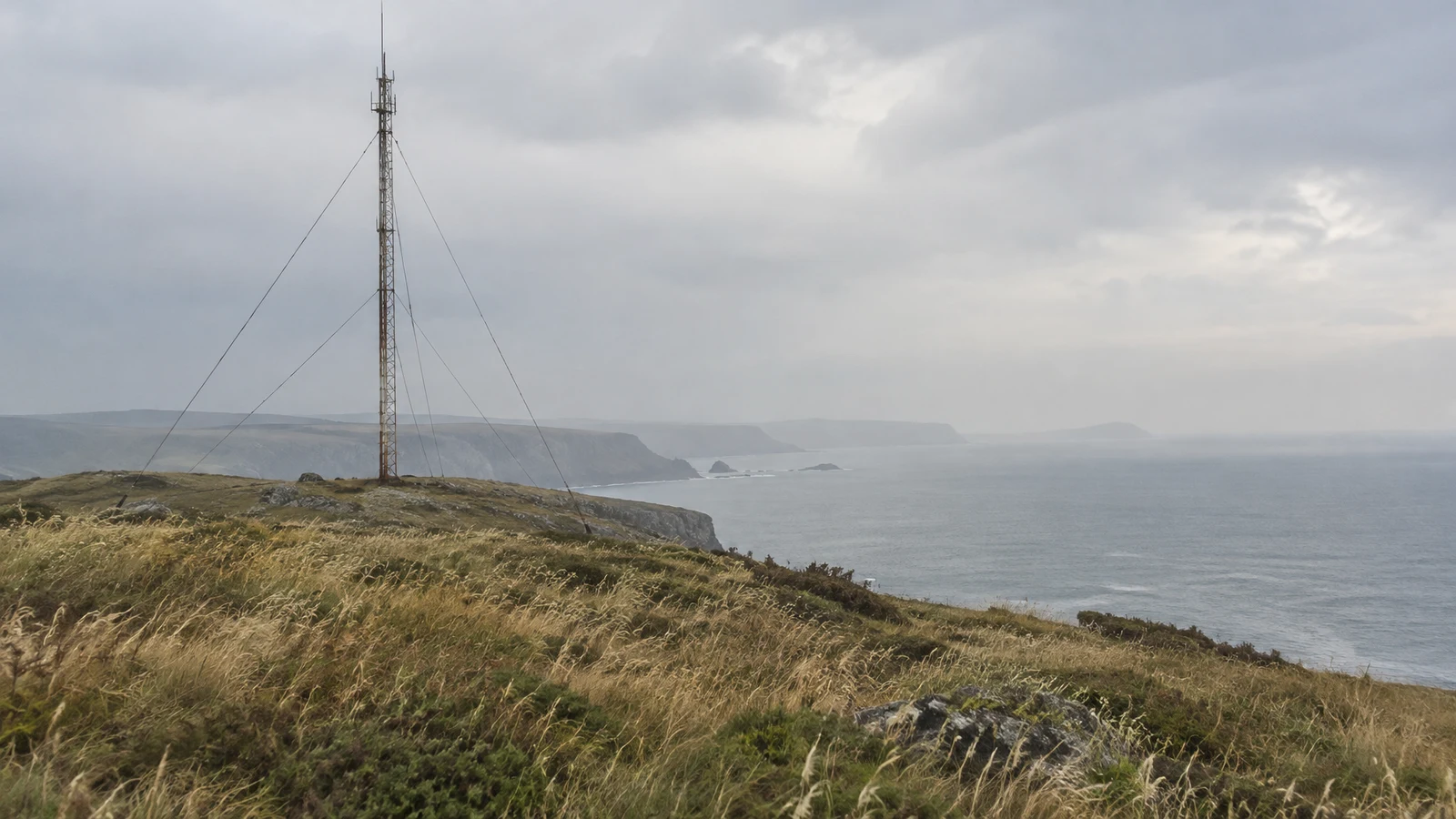 A weathered lattice radio mast with thin guy wires stands on a grassy coastal headland above a hazy grey sea, with distant cliffs beneath a broad overcast sky.