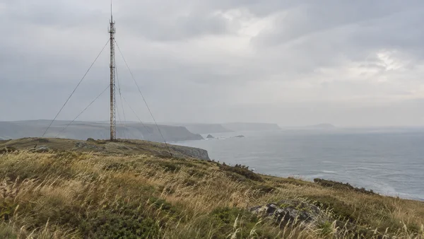 A weathered lattice radio mast with thin guy wires stands on a grassy coastal headland above a hazy grey sea, with distant cliffs beneath a broad overcast sky.