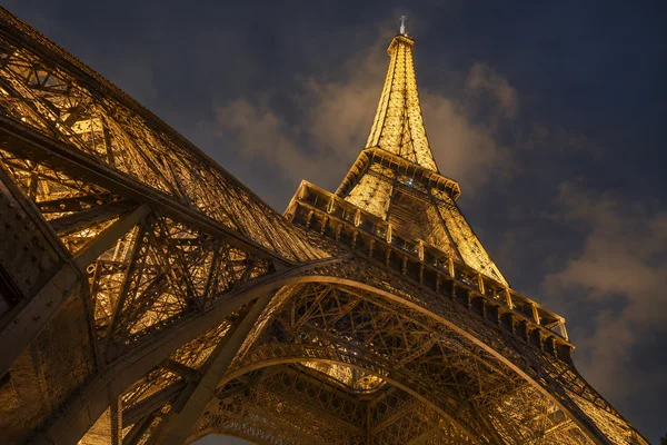 A steep upward view beneath the Eiffel Tower at night, with a massive illuminated iron leg filling the left foreground and the tower rising against a cloudy…