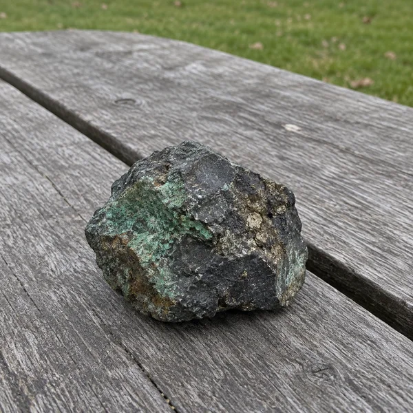 A rough dark rock with irregular green mineral patches and brassy flecks rests on a weathered grey wooden table, with a narrow strip of grass behind it.