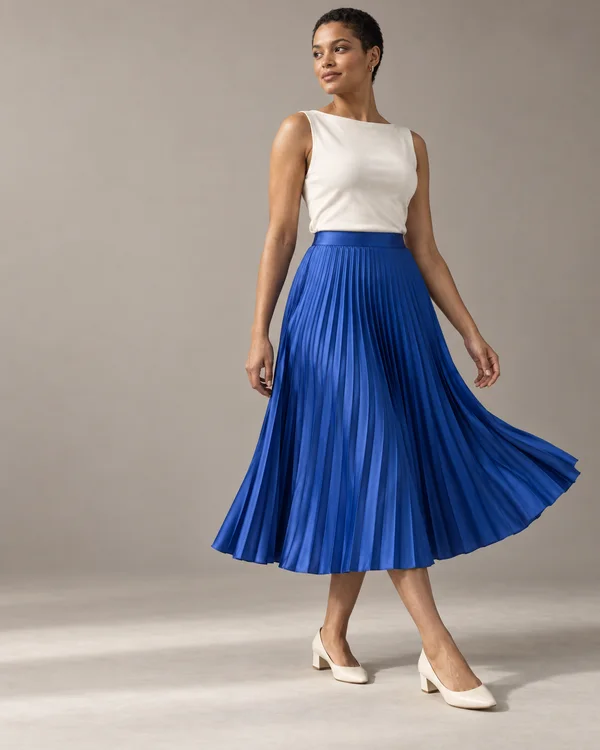 A woman with closely cropped dark hair wears an ivory sleeveless top, a cobalt-blue pleated midi skirt and ivory heels against a warm-grey studio backdrop.
