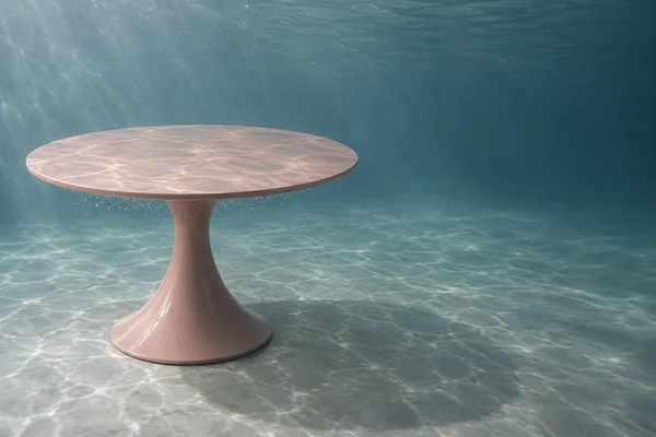 A blush-pink round pedestal dining table stands on a pale gray floor in a blue-green underwater setting, with rippling light patterns across its top and the…