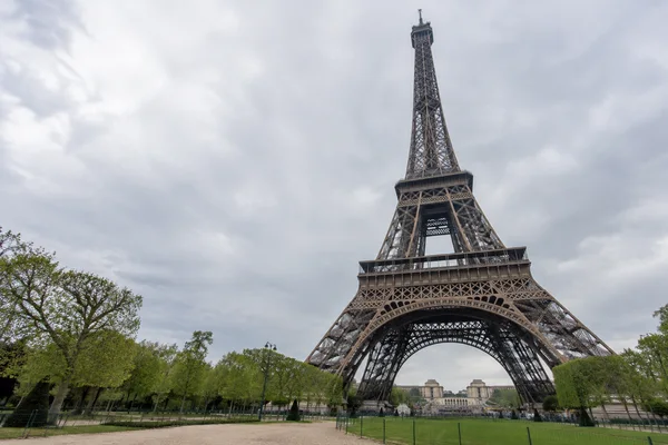 A low-angle view of the Eiffel Tower rising above trees, lawn and a gravel path beneath a pale, cloudy sky.