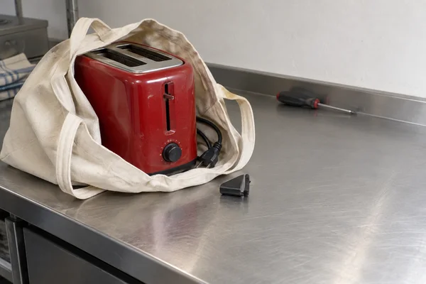 A scuffed red two-slot toaster sits inside an open cream canvas bag on a brushed metal counter.