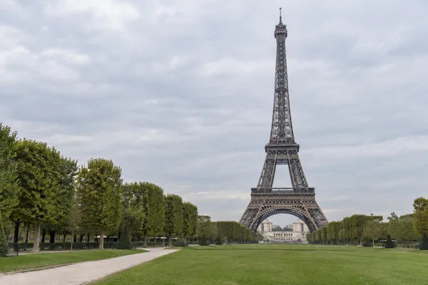 The Eiffel Tower stands slightly right of centre beneath an overcast sky, with a pale gravel path crossing the green lawn from the lower left and rows of trees…