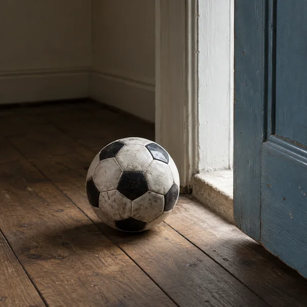 A scuffed black-and-white football rests on worn wooden floorboards beside a pale threshold inside a dim room.