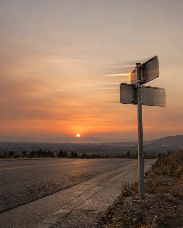 A blank two-blade metal street sign stands beside an empty paved road at sunset, with horizontal motion trails extending from the sign and a hazy cityscape…