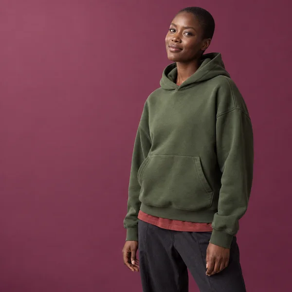 A dark-skinned woman with closely cropped hair models a moss-green hoodie over a brick-red shirt and charcoal trousers against a muted magenta studio backdrop…