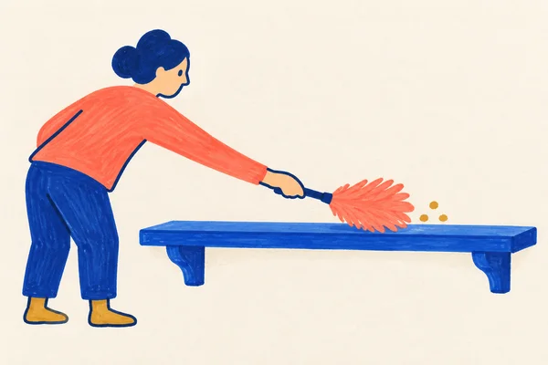 A person in a coral top bends from the left to dust a long cobalt-blue wall shelf with a coral feather duster.