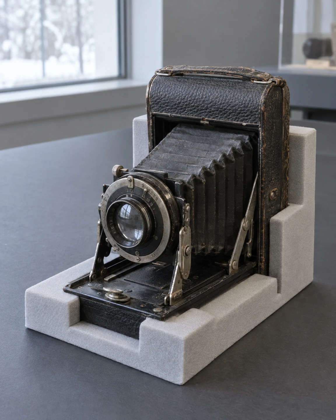 A worn vintage folding film camera with extended bellows and a circular lens sits in a pale grey foam cradle on a charcoal worktable, with a snowy window…
