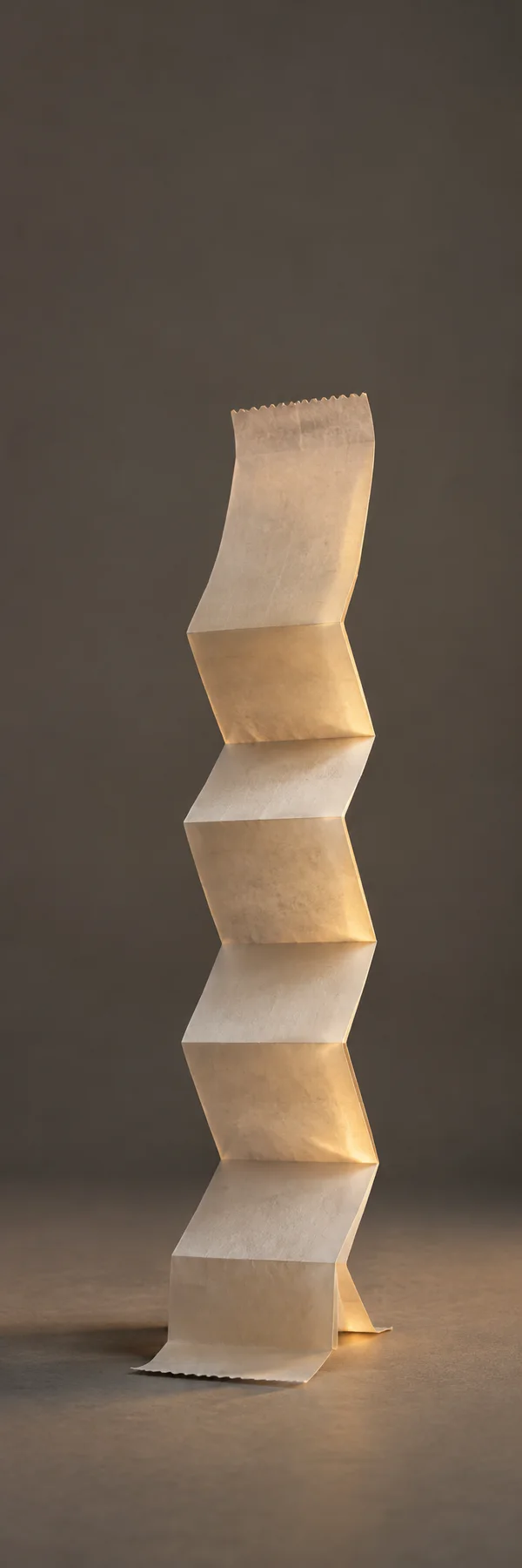 A long, blank, ivory-colored paper strip stands upright in broad alternating folds on a slate-colored studio floor, lit with warm amber light against a dark…