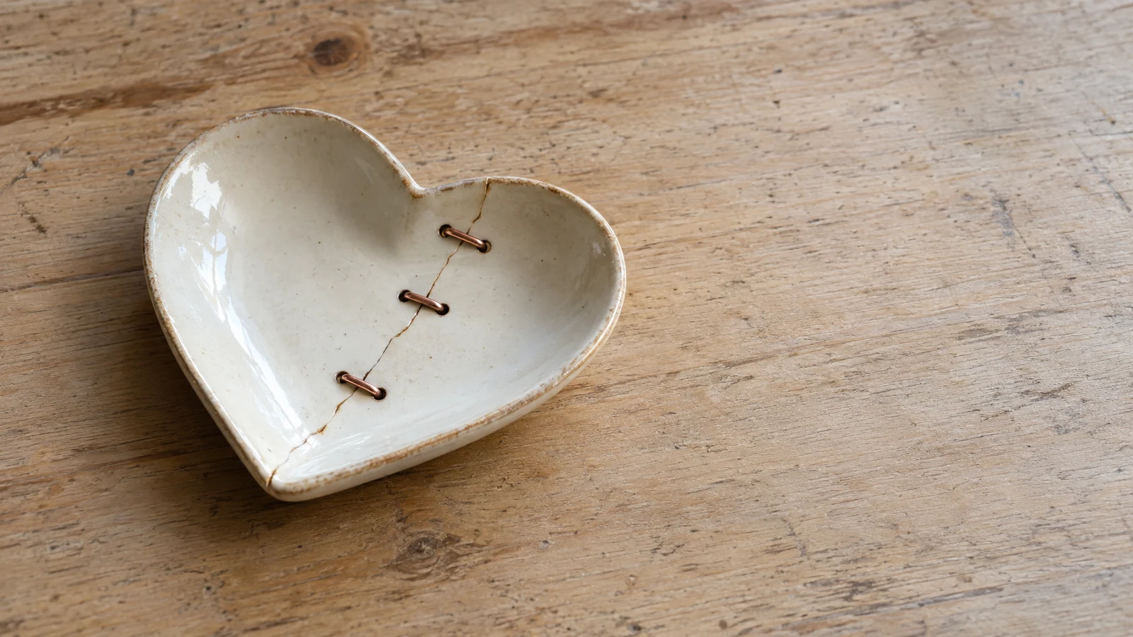 An ivory-glazed, heart-shaped ceramic dish with a diagonal crack secured by three copper staples rests left of center on a worn pale wooden table, with open…