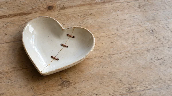 An ivory-glazed, heart-shaped ceramic dish with a diagonal crack secured by three copper staples rests left of center on a worn pale wooden table, with open…