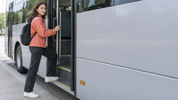 A young woman in a coral jacket and dark trousers boards a silver city bus, holding a vertical rail with one foot on the first step.