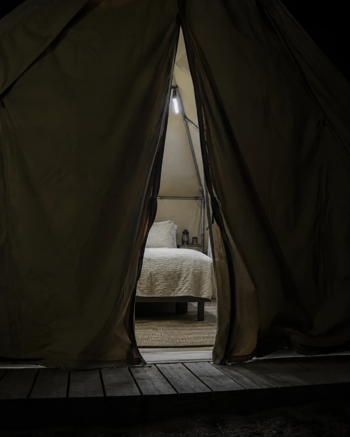 A partly open dark canvas glamping tent frames a raised bed with an ivory quilt, a small glowing LED batten, lanterns and a rug; timber deck boards cross the…