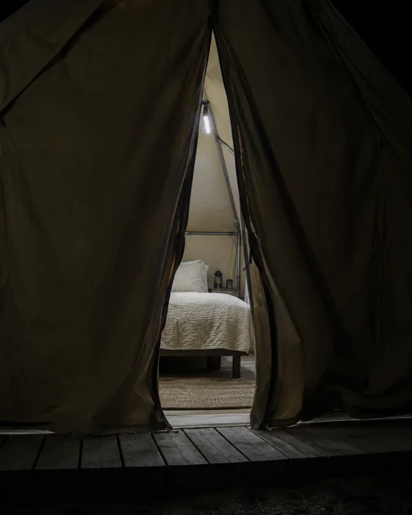 A partly open dark canvas glamping tent frames a raised bed with an ivory quilt, a small glowing LED batten, lanterns and a rug; timber deck boards cross the…